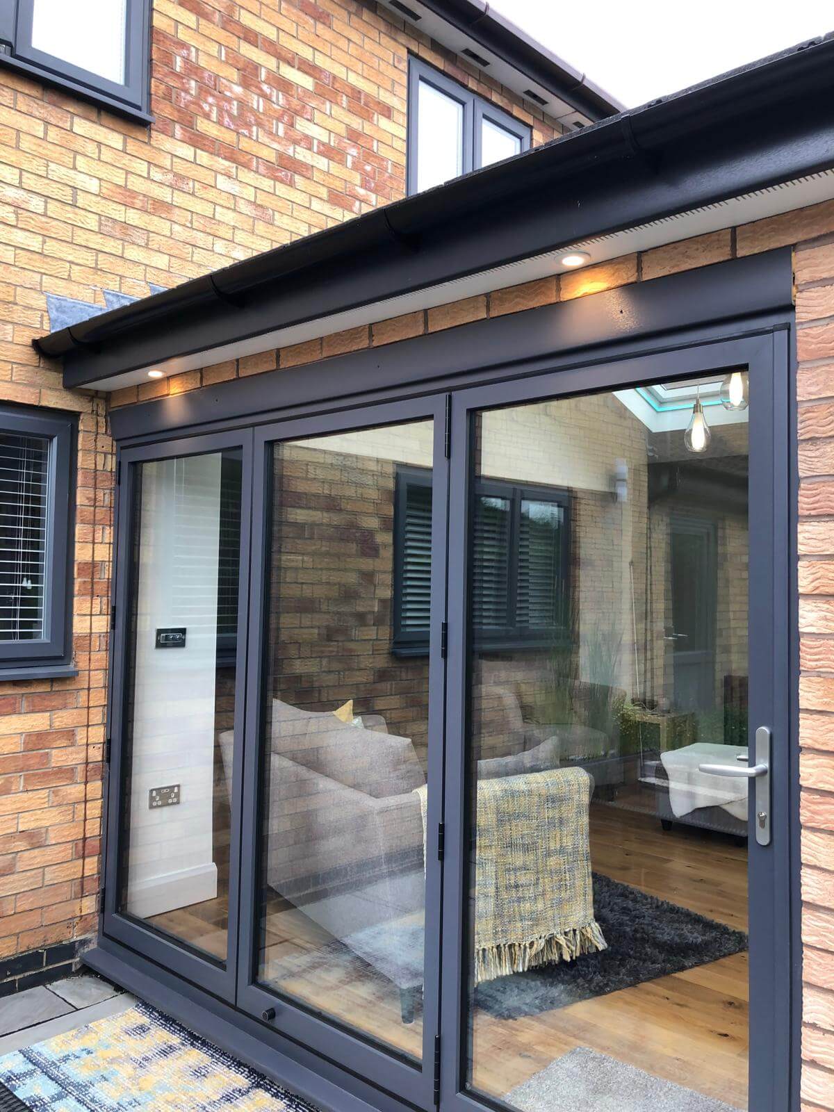 Grey bifold doors on a brick exterior, reflecting a modern living room with a sofa and patterned rug.